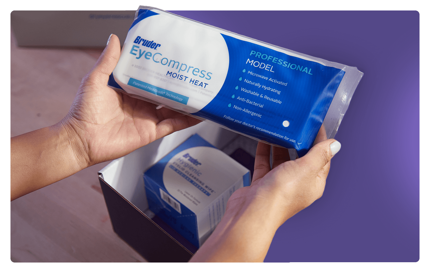Hands holding a Bruder EyeCompress Moist Heat pack against a purple background. Another pack is visible inside an open box on a wooden surface. The packaging highlights features such as reusable and anti-bacterial.