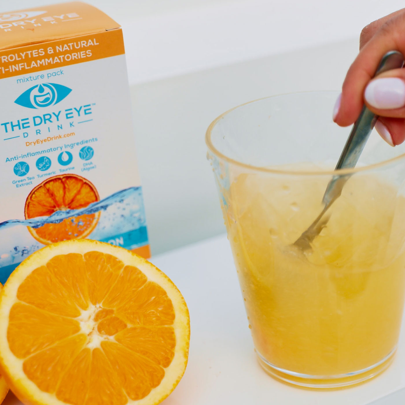 A person is stirring a yellow drink in a clear glass with a spoon. A box labeled The Dry Eye Drink and a sliced orange are on the table. The box mentions electrolytes and natural anti-inflammatories.