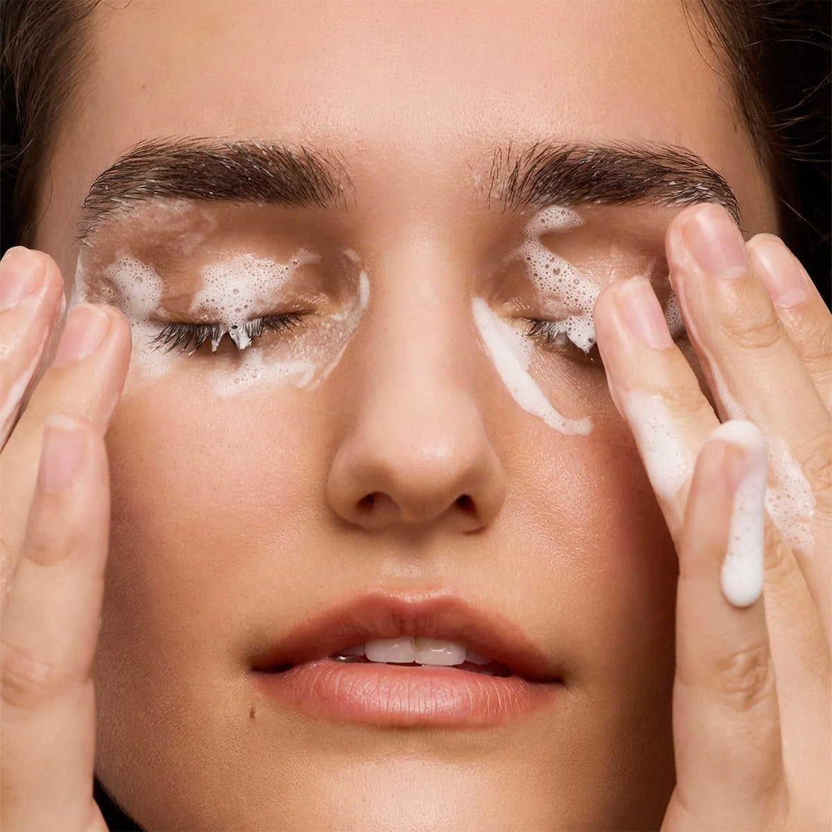Using the Daily Practice Purifying Foam Cleanser – Gentle & Hydrating for Eyes & Lashes (80mL), a person cleanses their closed eyes, keeping fingers near and foam visible on their skin, highlighting a gentle skincare routine focused on eye area protection.