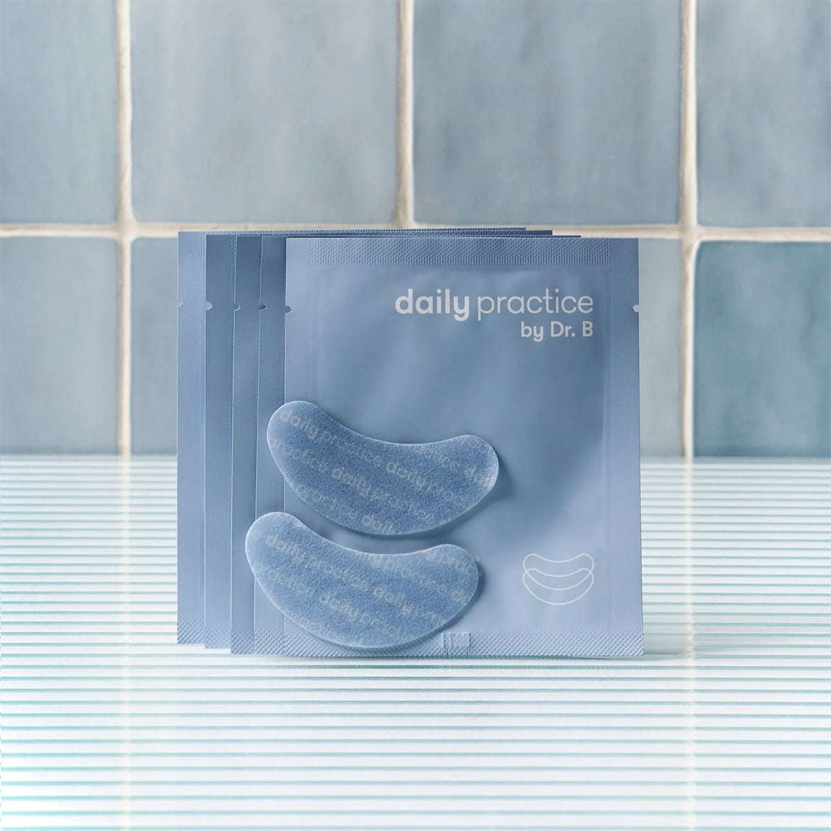 Four packets of Daily Practice Brighten & Hydrate Eye Patches sit on a striped surface against a tiled backdrop. Two crescent-shaped patches, meant to refresh and rejuvenate, lie outside one packet. The color scheme is blue and white.