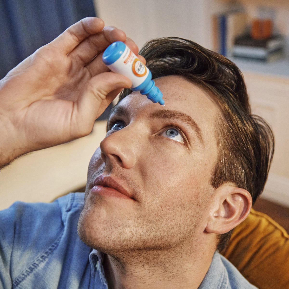 Indoors with blurry books and curtains in the backdrop, a person in a denim shirt tilts their head back to apply Bausch + Lomb Blink Triple Care 3 in 1 Eye Drops into their left eye for long-lasting dry eye relief.