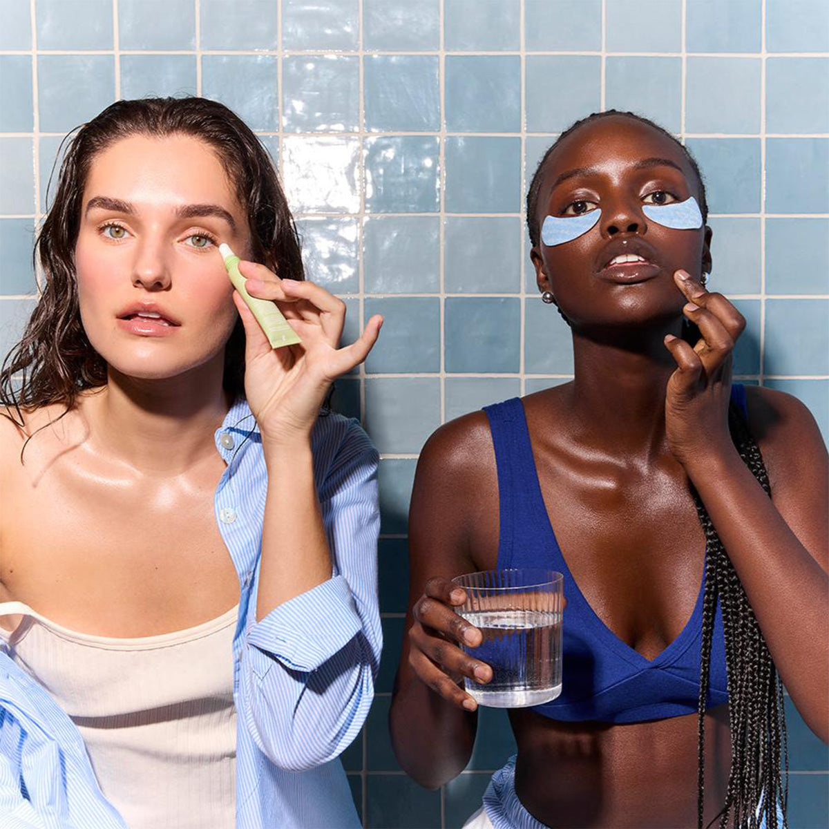 In a blue-tiled bathroom, two women with wet-look hairstyles and casual tops tend to their skincare. One applies Daily Practice Firm & Restore Eye Cream, known for its smoothing and hydrating properties, while the other uses eye patches to enhance skin elasticity and holds a glass of water.