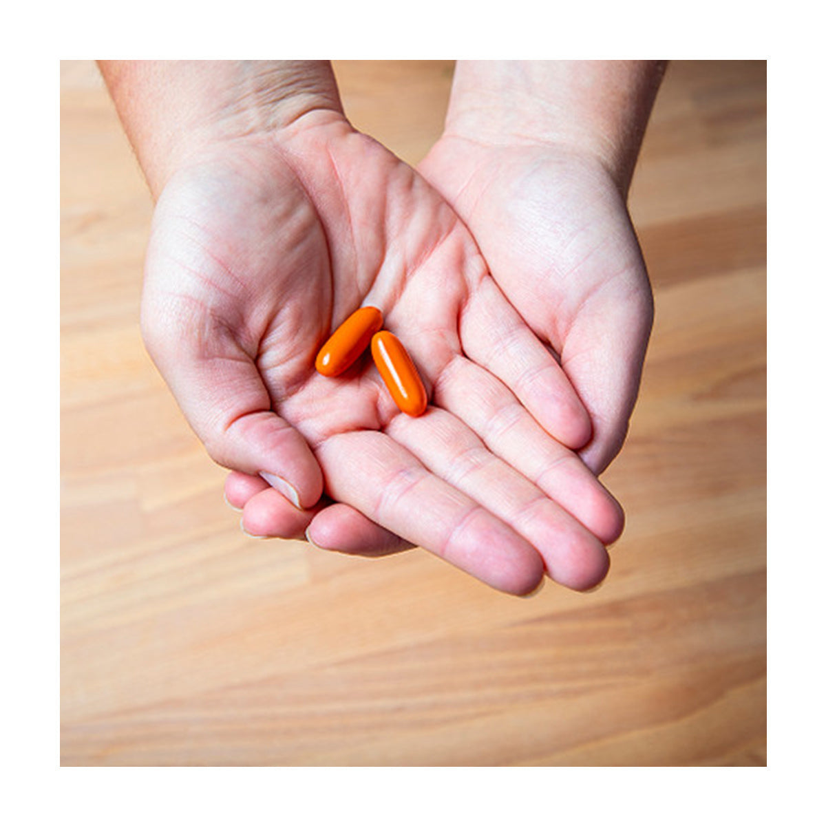 Hands cupped together hold two orange capsules, likely EyePromise® EZ Tears – Natural Dry Eye Relief with Omega-3s, Turmeric, and Green Tea Extract (60ct), set against a wooden floor background.