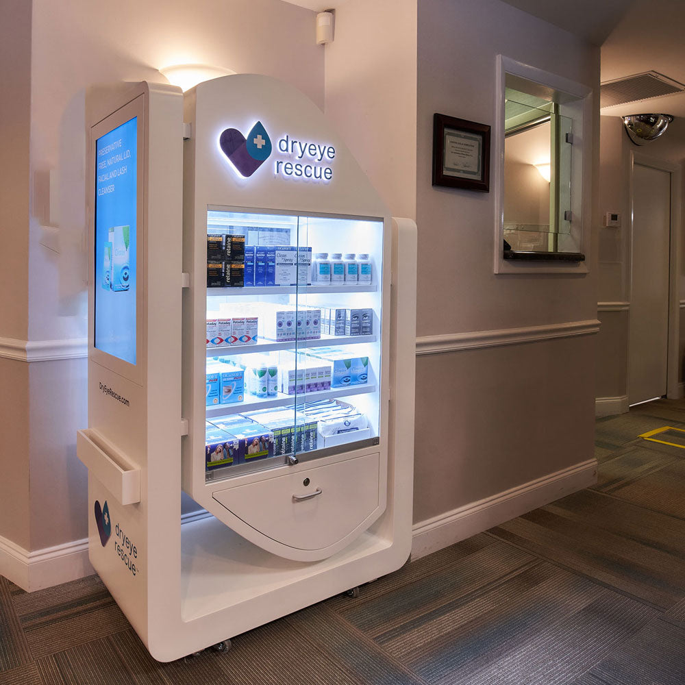 A DryEye Rescue vending machine titled Free Standing Floor Display Free Rent (Large) with a digital screen stands in a well-lit hallway. The walls are light-colored, and a framed certificate is beside a window to the right. It dispenses various eye care products.