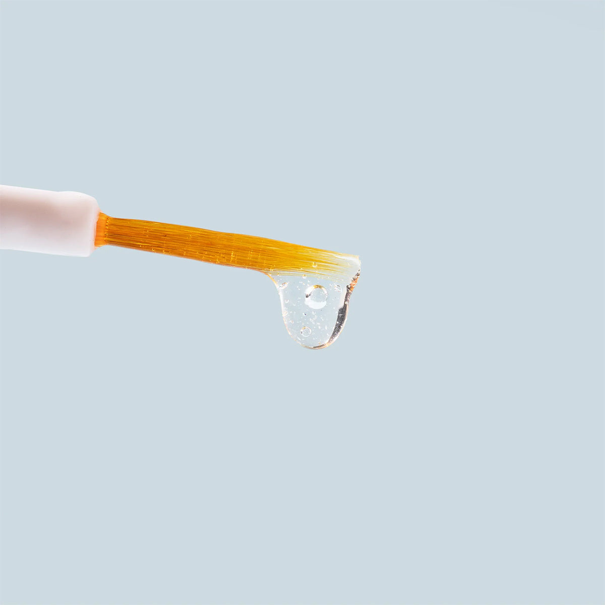 Close-up of Daily Practice Lash Enhancing Serums orange-handled brush with white bristles, holding a drop of the clear lash serum against a plain light blue background, ideal for naturally achieving fuller, longer, healthier lashes.