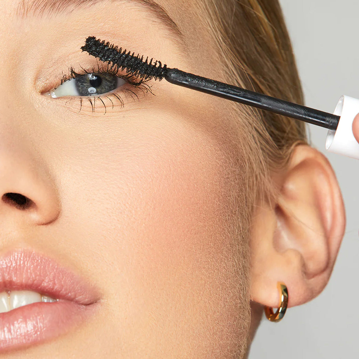 A close-up of someone applying Twenty Twentys Clean Sweep Mascara in Pitch Black shows a black brush near the lashes. The ophthalmologist-approved formula is safe for contact lens wearers. The person, with light skin, full lips, and a small hoop earring, gazes upward as they apply mascara.