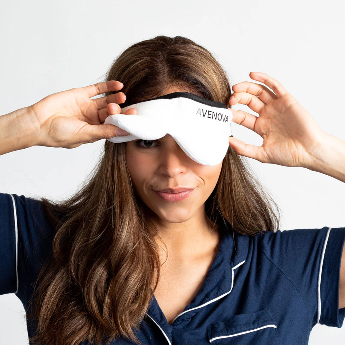 In a plain white setting, someone with long hair in navy pajamas adjusts a NovaBay Avenova Microwavable Heat Eye Compress Mask over their eyes, ideal for blepharitis relief.