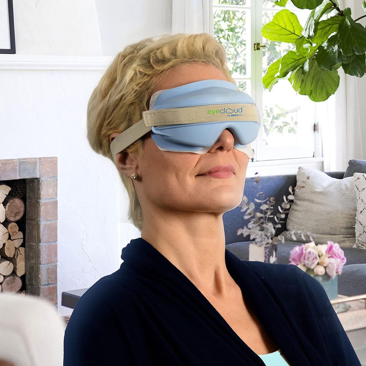 A person with short blond hair relaxes in a cozy living room, wearing the PRN EyeCloud Home Treatment Kits blue eye mask. The warm ambiance from the fireplace, sofa, and floral decor complements their soothing dry eye relief experience as they enjoy moist heat therapy with 600+ gel pack uses.