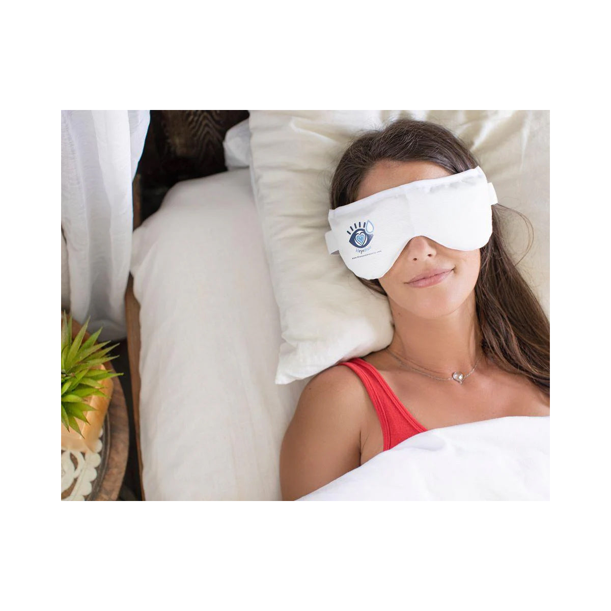 A woman in a red tank top lies on a bed, covered with a white blanket and wearing the Heyedrate Warm Compress Eye Mask from PRN, featuring a blue eye design for dry eyes relief. Beside her is a wooden table with a potted plant.