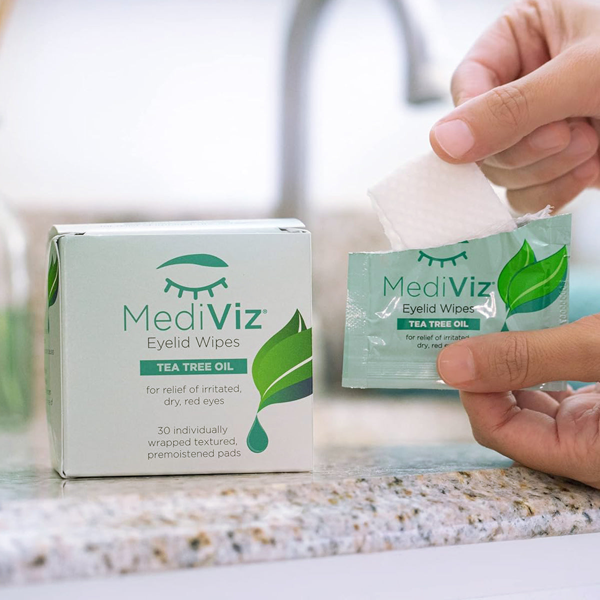 A person holds an opened packet of green and white Mediviz Tea Tree Eyelid Cleansing Wipes by PRN on a countertop, with the box stating they relieve irritated, dry, and red eyes.