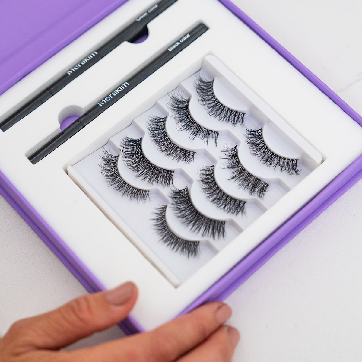 A hand holds a purple Merakim Eyes on Beauty Kit box with faux mink lashes and two black adhesive/eyeliner pens. The open box displays the contents neatly arranged on a white background.
