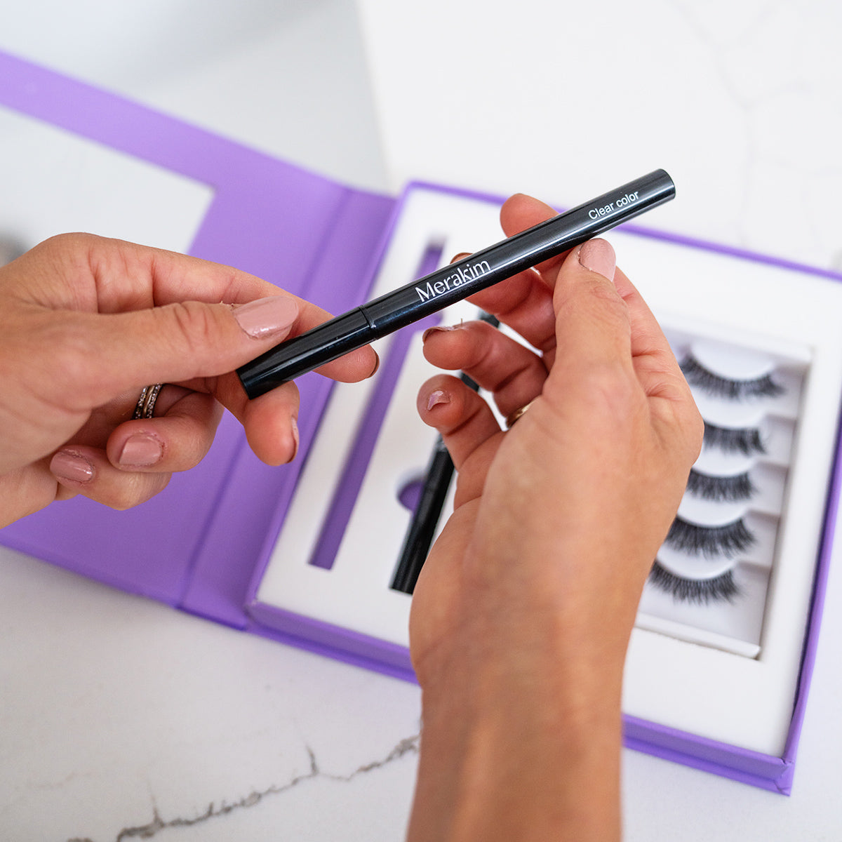 A person holds a Merakin clear glue pen while a box of Merakim Eyes on Beauty Lashes, an alternative to eyelash extensions, is open in the background. The purple box sits on a white surface alongside faux mink lashes (5 pairs).