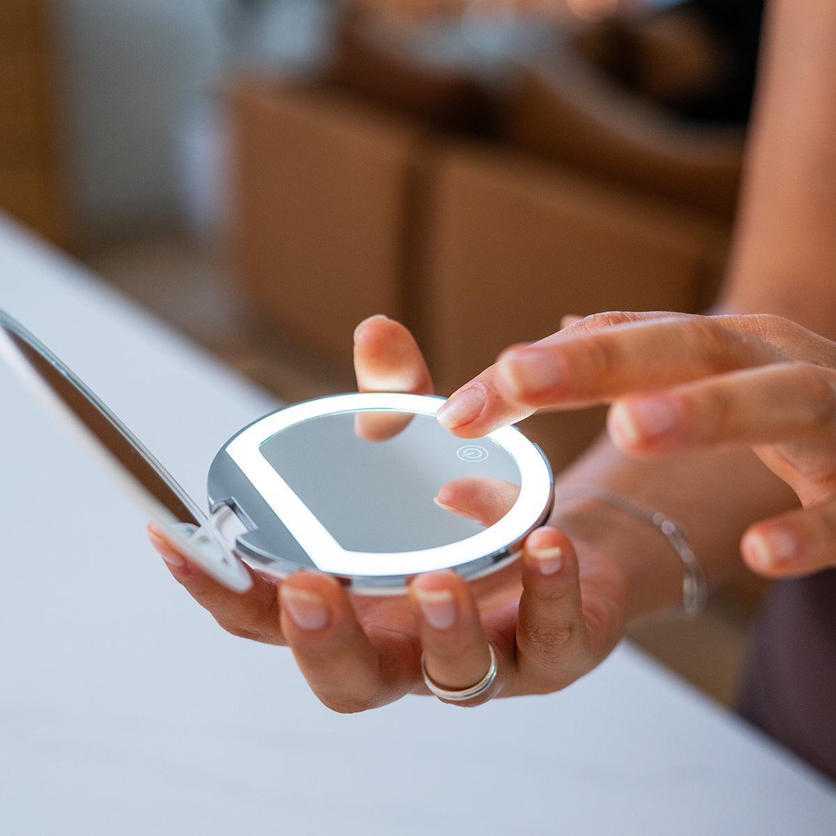A person gently touches a Merakim Rechargeable LED Makeup Mirror with 1/3X Magnification. They wear a bracelet on their right wrist, and the blurred background enhances the focus on the mirrors sleek design, adjustable light settings, and eco-friendly portability.