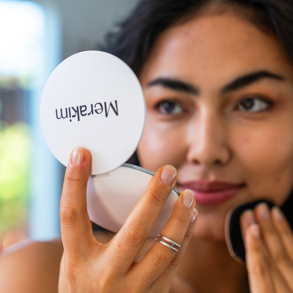 A person uses the Merakim Rechargeable LED Makeup Mirror with 1/3X magnification and adjustable light settings to apply makeup, holding the white compact mirror labeled Merakim against a softly blurred background for an enhanced makeup application experience.