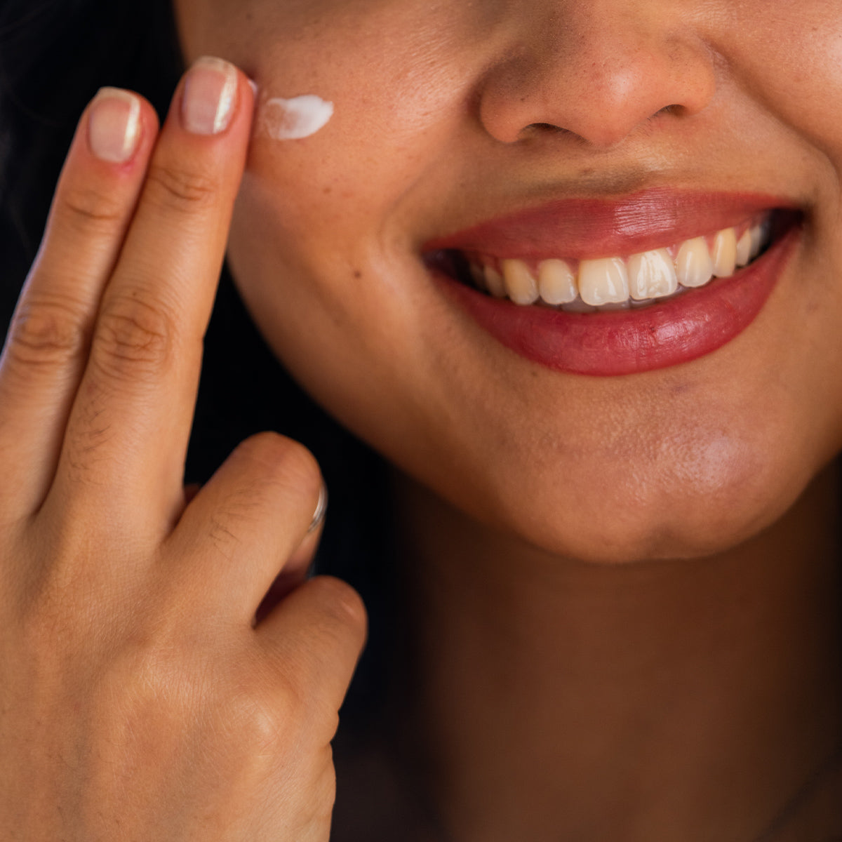 A person with manicured nails and light polish smiles brightly while applying Merakim Facial Sunscreen SPF 50 (50mL) to their cheek with two fingers, showcasing a joyful expression.