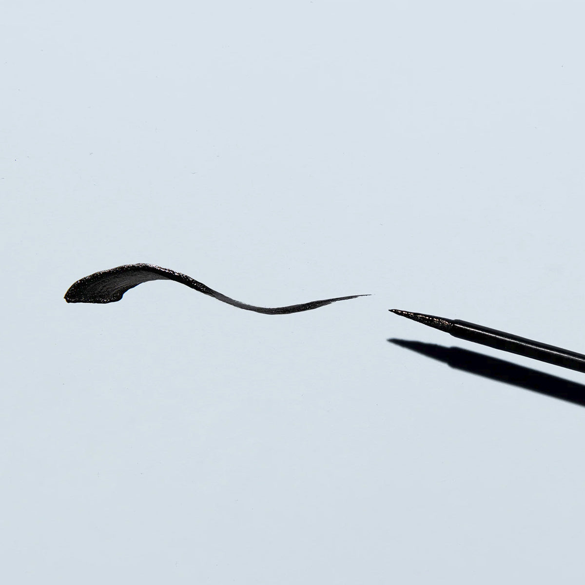 The Twenty Twenty Double Duty Precision Eyeliner in Pitch Black, enriched with eye-safe ingredients, creates a smooth, curved line. The black liquid brush casts a soft shadow on a light gray background as it is positioned to the right.