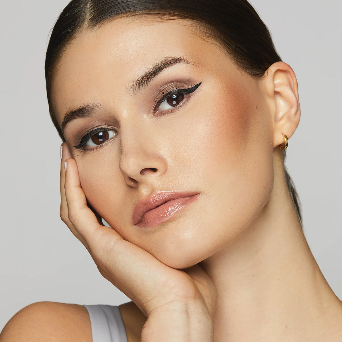 A person with sleek dark hair and winged eyeliner rests their face on their hand, wearing a small hoop earring and a light-colored top. They embody eye-safe beauty using the Twenty Twenty Beauty Starter Set: Facial Spray, Eyeshadow Sticks, Lash & Brow Serum & Mascara, gazing softly at the camera against a plain backdrop.