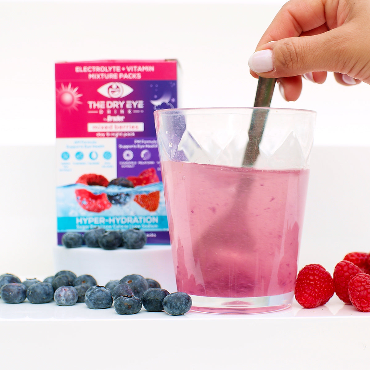 A hand stirs a glass of pink liquid with a spoon, promoting hydration for dry eye relief. In the background, there’s a box labeled Bruder Dry Eye Drink™ Complete Set, featuring blueberries and raspberries. Fresh berries surround the glass, enhancing the appeal of this refreshing hydration solution.