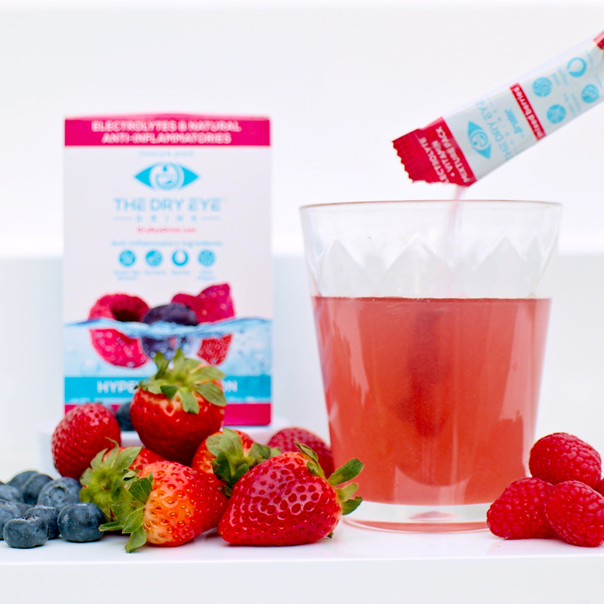 A glass of pink beverage gets a sachet pour from Bruders Dry Eye Drink™ Daytime. Fresh strawberries and blueberries surround the glass, while a background box labeled Dry Eye Drink™ with vibrant fruit images highlights this doctor-formulated electrolyte solution for eye health.