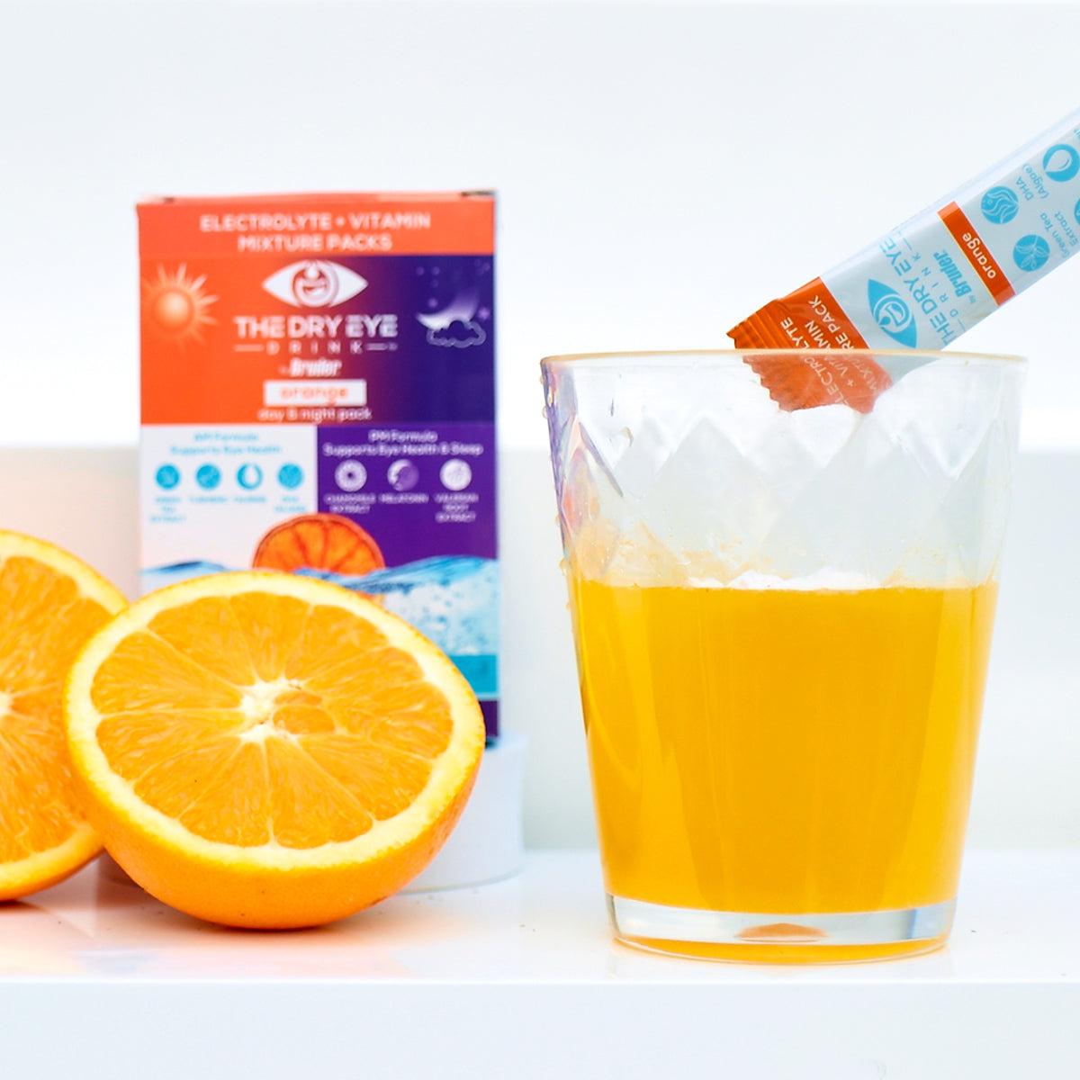 A glass with an orange-flavored drink and a halved orange sits alongside a sachet labeled Dry Eye Drink™ Complete Set, from Bruder, promoting hydration for dry eyes. In the background, its box highlights electrolytes and vitamins designed for both daytime and nighttime use.