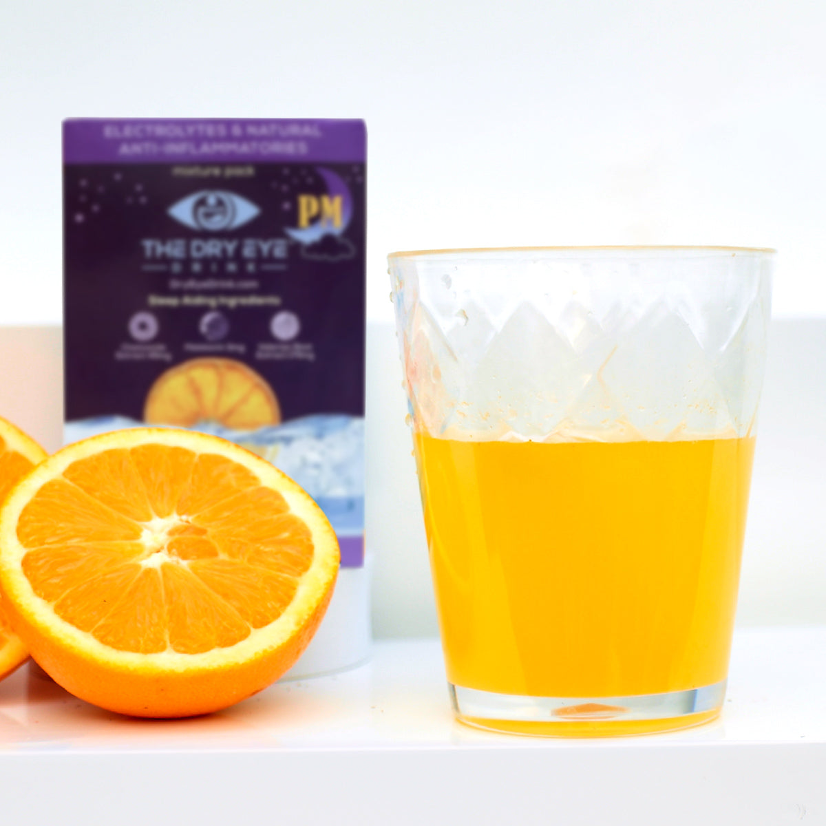 A glass of orange juice is in the foreground on a white surface. Sliced oranges sit invitingly to the left. In the background, a purple box labeled Dry Eye Drink™ Daytime by Bruder displays images of an orange and ice, highlighting promises of dry eye relief and improved eye health.