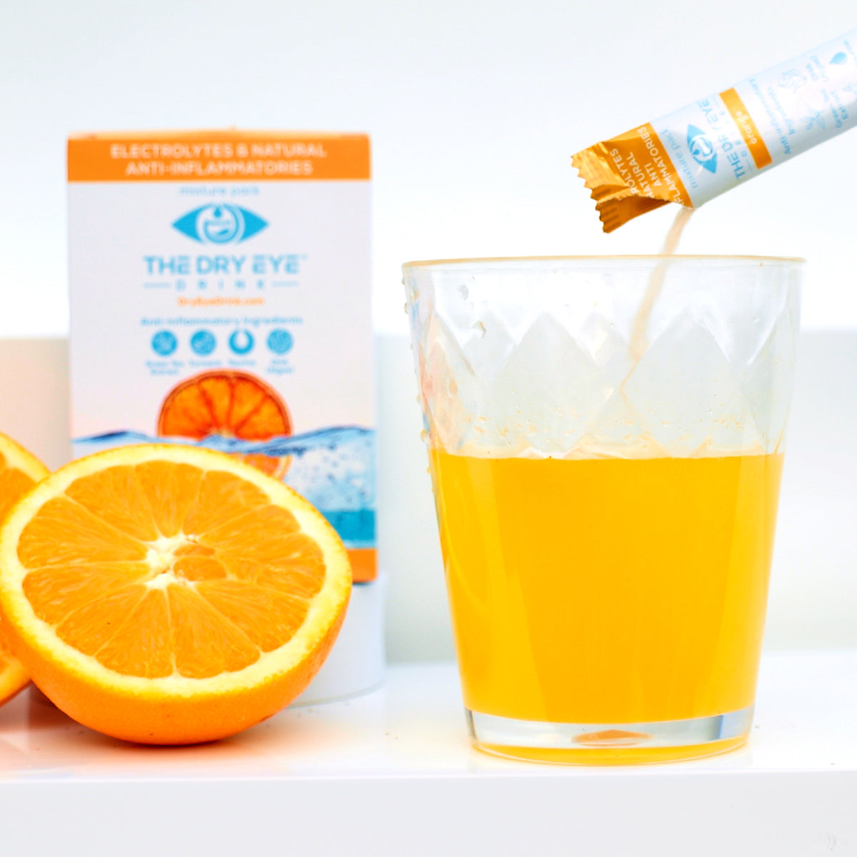 A glass of orange liquid with powdered contents from a sachet is in the foreground, flanked by two orange halves. In the background, a Bruder box labeled Dry Eye Drink™ Daytime offers doctor-formulated electrolyte hydration for eye health with natural anti-inflammatories.