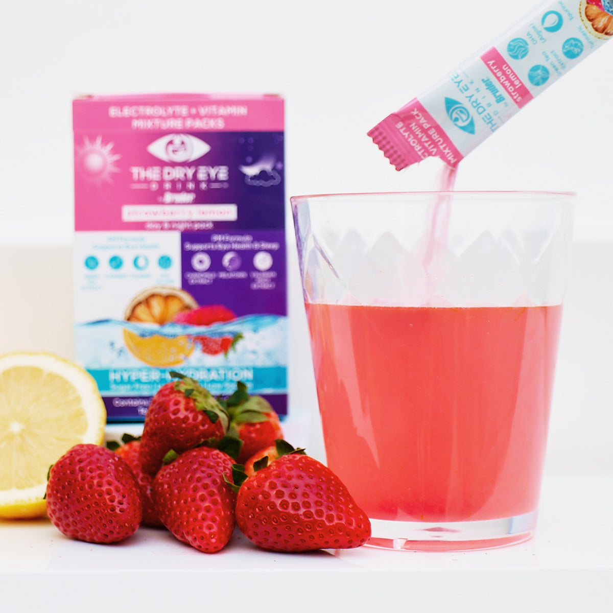 A glass of pink drink with fresh strawberries and a lemon slice beside it is being mixed with Bruders Dry Eye Drink™ Complete Set – Daytime & PM Hydration for Dry Eyes (Strawberry Lemonade). A vibrant pink and purple product box is seen in the background, highlighting hydration benefits.