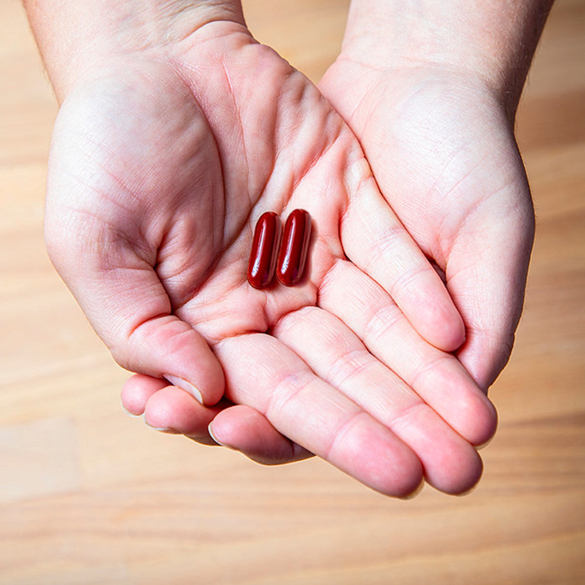 Hands gently cradle two red capsules of EyePromise Macular Health Complete AREDS 2 based Multivitamin (60ct) against a serene wooden floor backdrop.