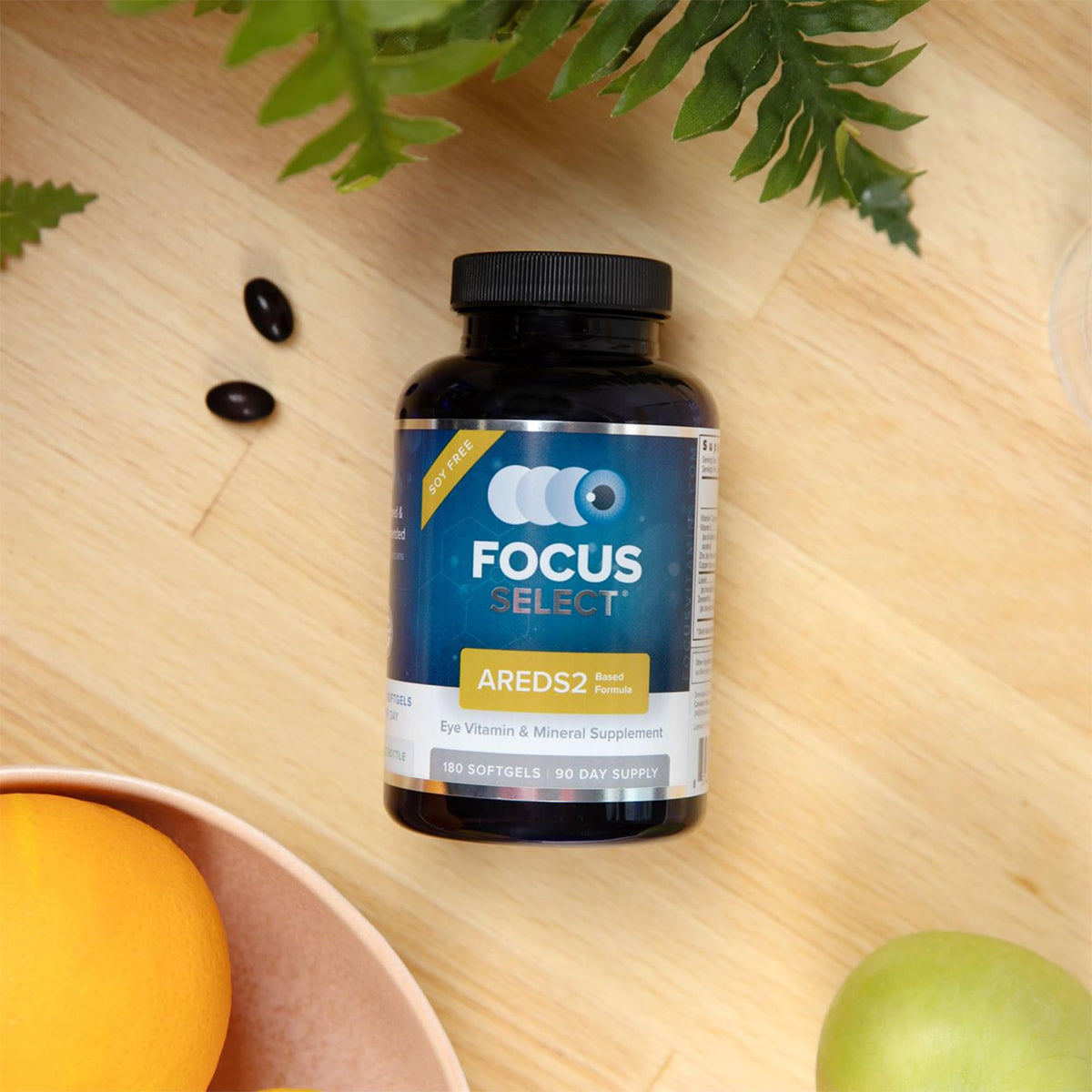 A Focus Select Soy Free Supplement bottle from Focus Vitamins, featuring an AREDS2 formula for macular degeneration (180ct. 90 Day Supply), is placed on a light wooden surface with green leaves, two black softgels, an orange in a wooden bowl, and a green apple around it.