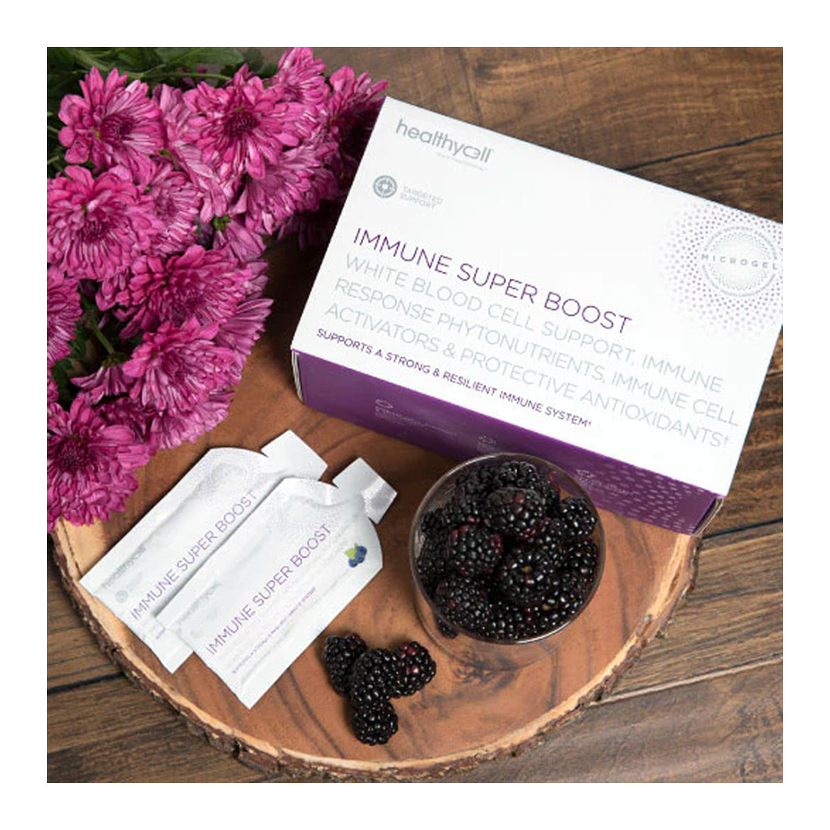 A HealthyCell Immune Support Boost Gel Pack box sits on wood, with two sachets and a glass bowl of blackberries. Purple flowers adorn the left side of this Echinacea and Elderberry display.