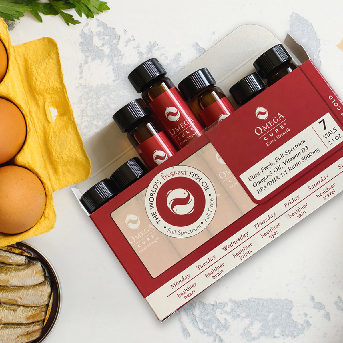 A box of Omega Cure Extra Strength Fresh Liquid Omega from Omega 3 Innovations is displayed on a table with brown eggs, lemon peel, and fish. The red and white packaging holds 7 vials for a weekly supply, highlighting freshness and full-spectrum benefits. Cold shipping included.