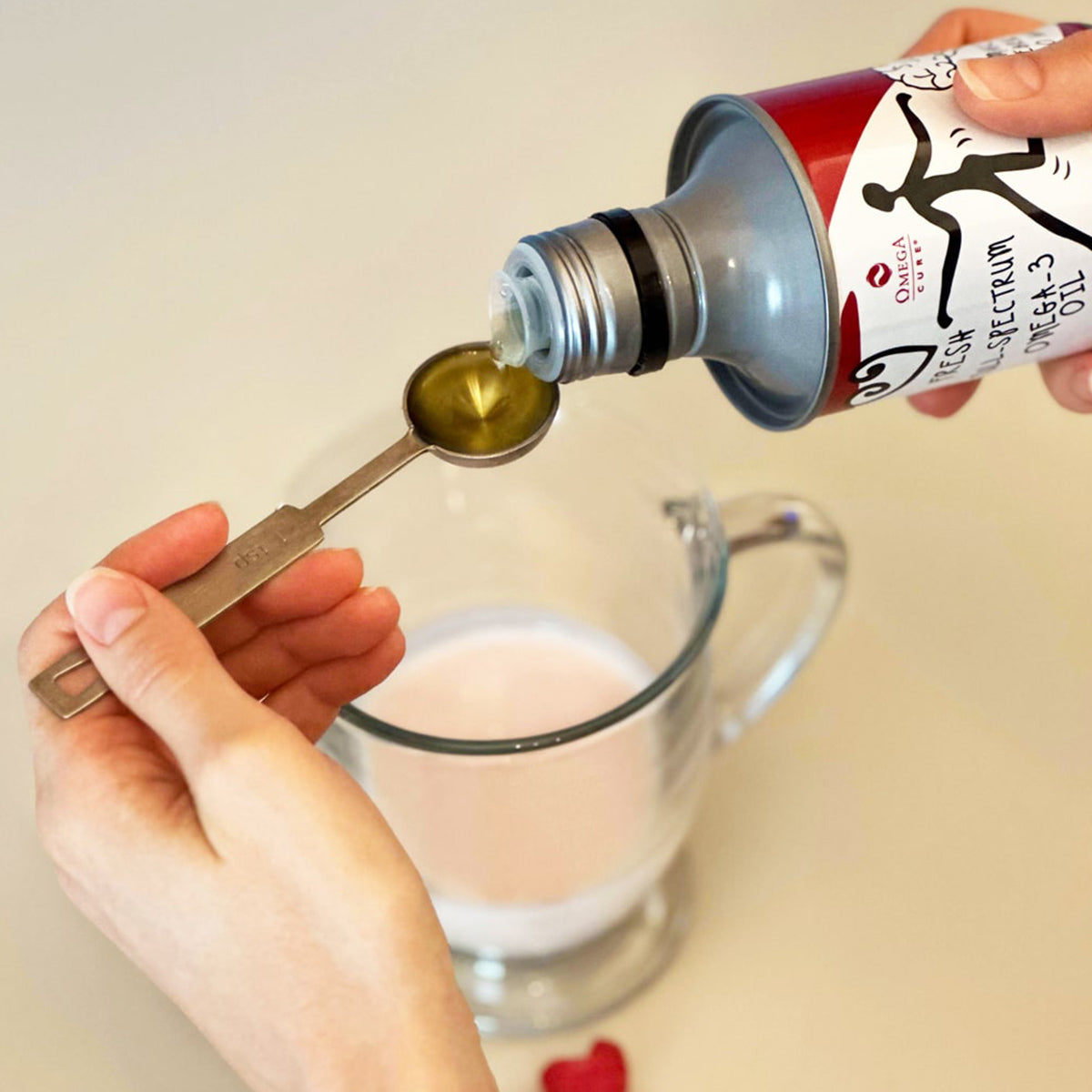 A person pours Omega Cure Omega 3 Fish Oil from an 8.5 oz (250mL) canister into a measuring spoon over a clear glass mug with pink liquid. The Omega 3 Innovations canister features black text and graphics. Known for its non-fishy taste, the oil pairs well with any drink. A small red item is on the table below the mug.