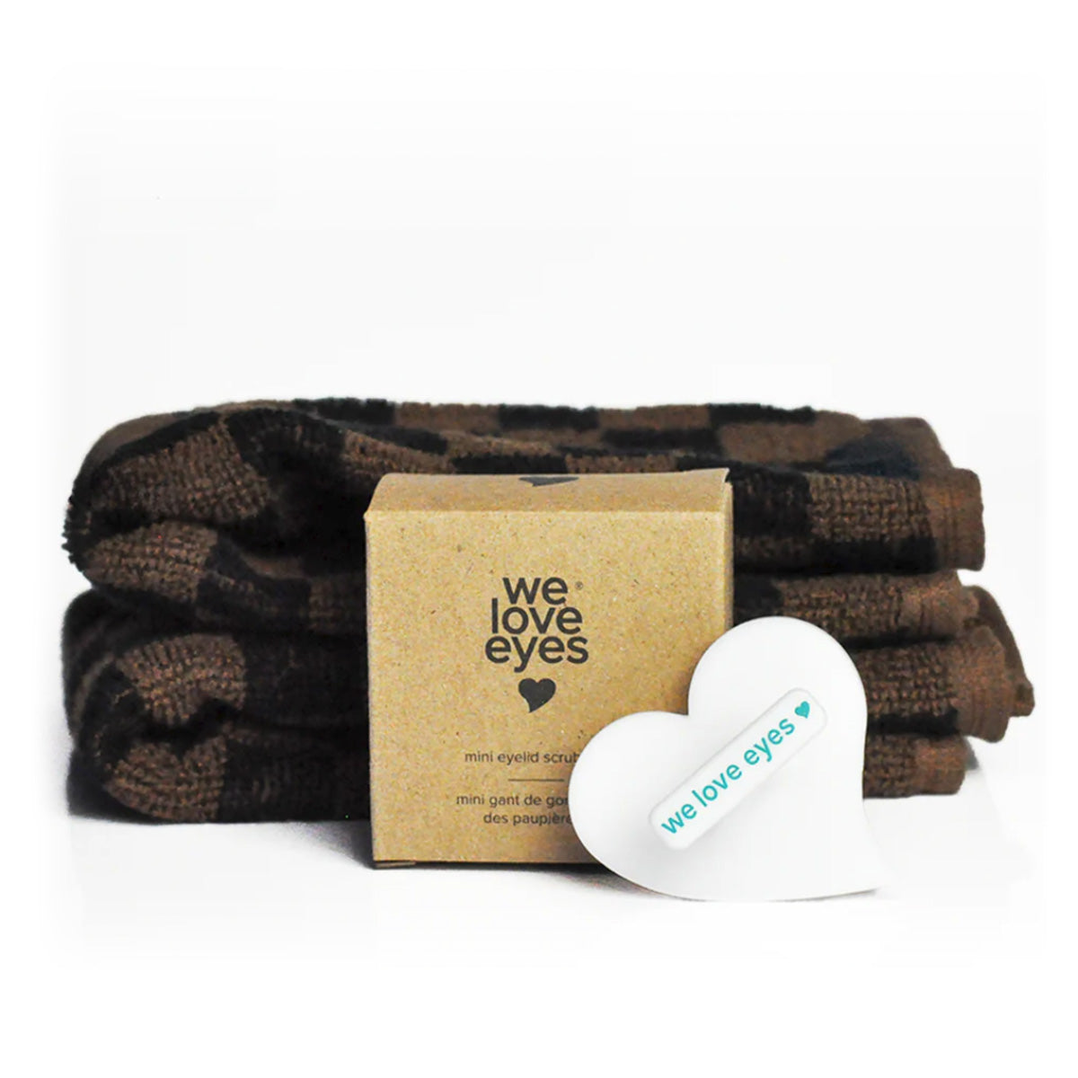 A stack of dark towels and a small We Love Eyes box sit on a white surface, with a heart-shaped item in front. The brand name and logo, featuring a small teal dot, highlight the We Love Eyes Mini Eyelid Scrub Mitt for precision cleansing and healthy lid hygiene.