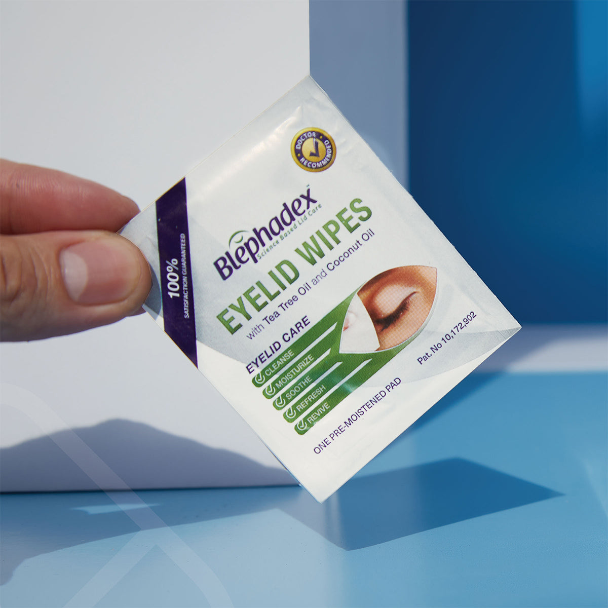 A hand holds a sachet labeled Blephadex Eyelid Wipes by Lunovus, featuring a closed eye image. The packaging promotes tea tree and coconut oil for 100% effective eyelid hygiene against dust and pollen. It offers eye itch relief in a 1-month box of 30 wipes.