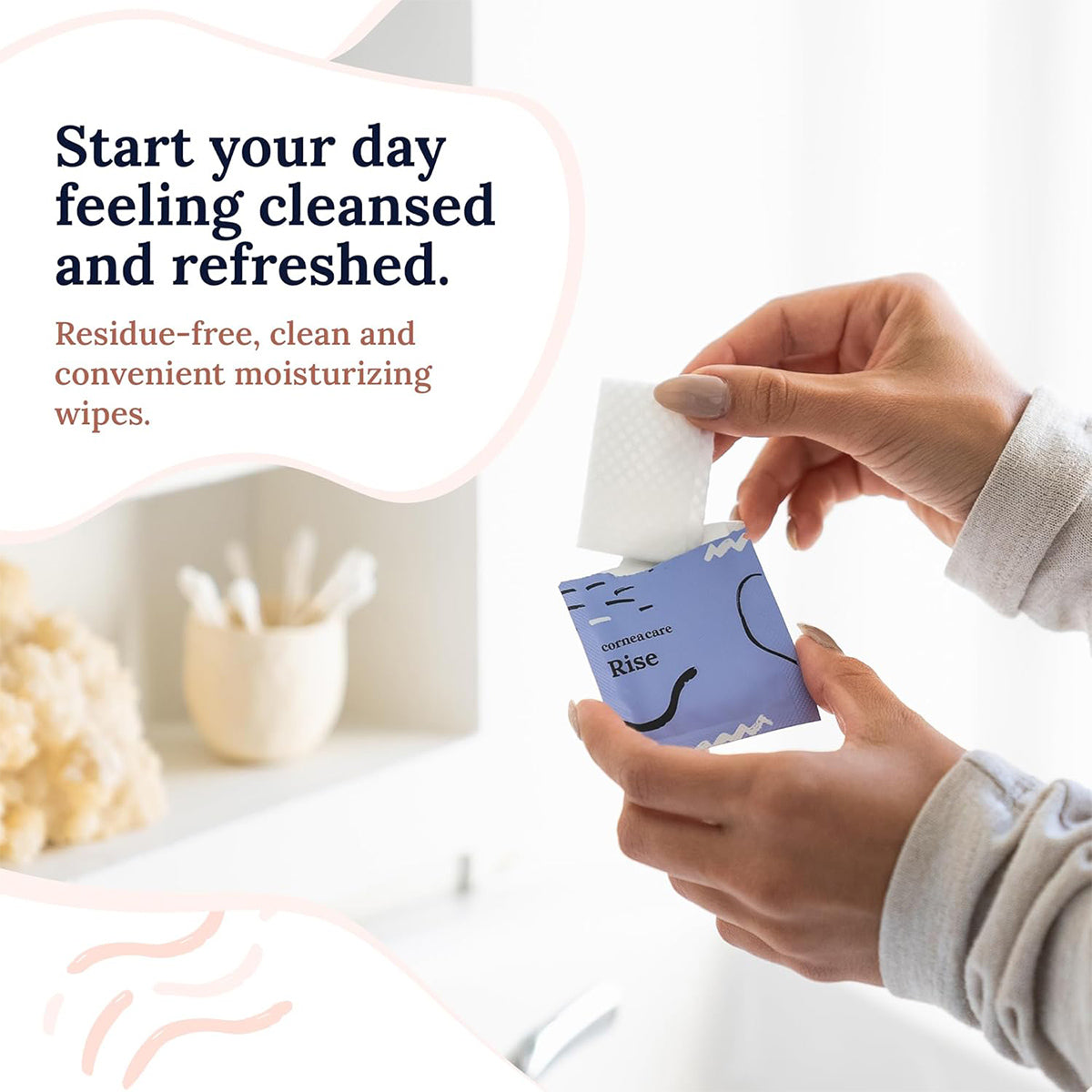 In a blurred bathroom background, someone holds a blue packet of CorneaCare Wipes with Tea Tree Oil (30ct), partially pulling one out. The text reads: Start your day feeling cleansed and refreshed with residue-free, convenient moisturizing wipes.
