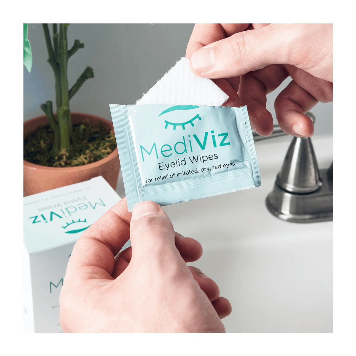 Hands open a MediViz Eyelid Cleansing Wipes packet by PRN - Physician Recommended Nutriceuticals. The light blue pack, featuring an eye illustration, alleviates dry, irritated eyes and supports eye health. A plant and silver faucet are softly blurred in the background.