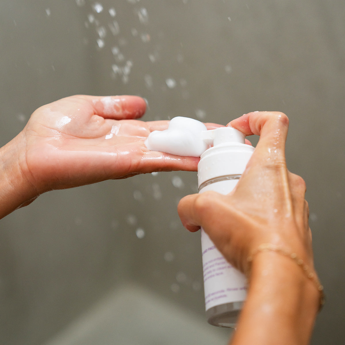 In a shower, a person dispenses Merakim Tea Tree Oil Foam Facial Cleanser (120mL) onto their palm under running water. The focus is on hands and bottle, with water droplets visible, emphasizing its soothing relief for Dry Eye Disease.