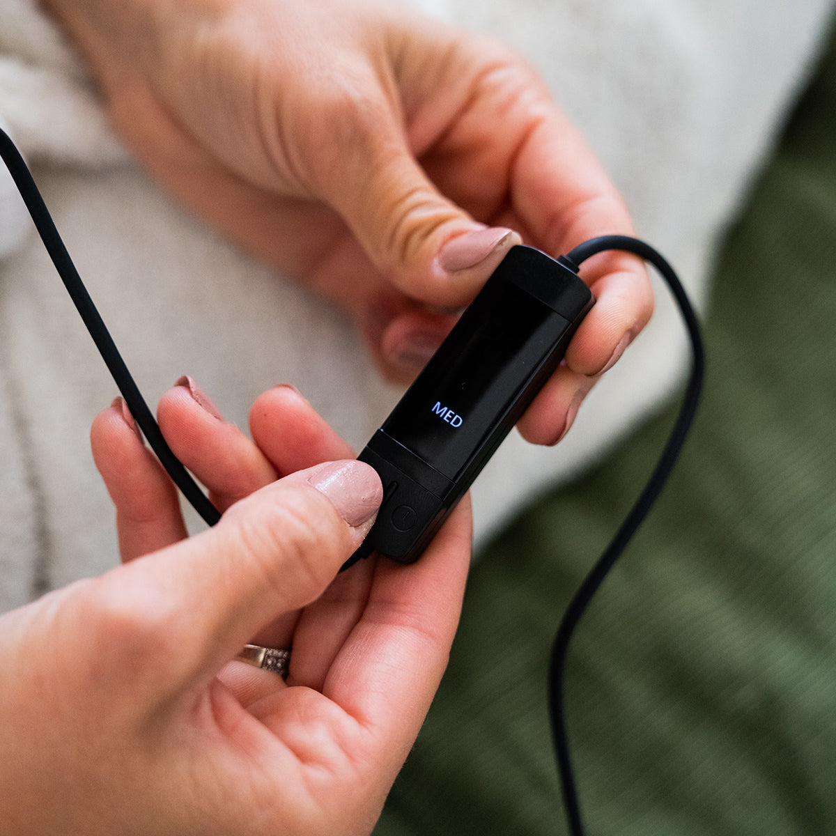 Close-up of hands holding the Merakim Professional USB Heat Eye Mask with a screen displaying MED. It connects via cable, and the person sports light nail polish and a silver ring. A textured fabric is in the background.