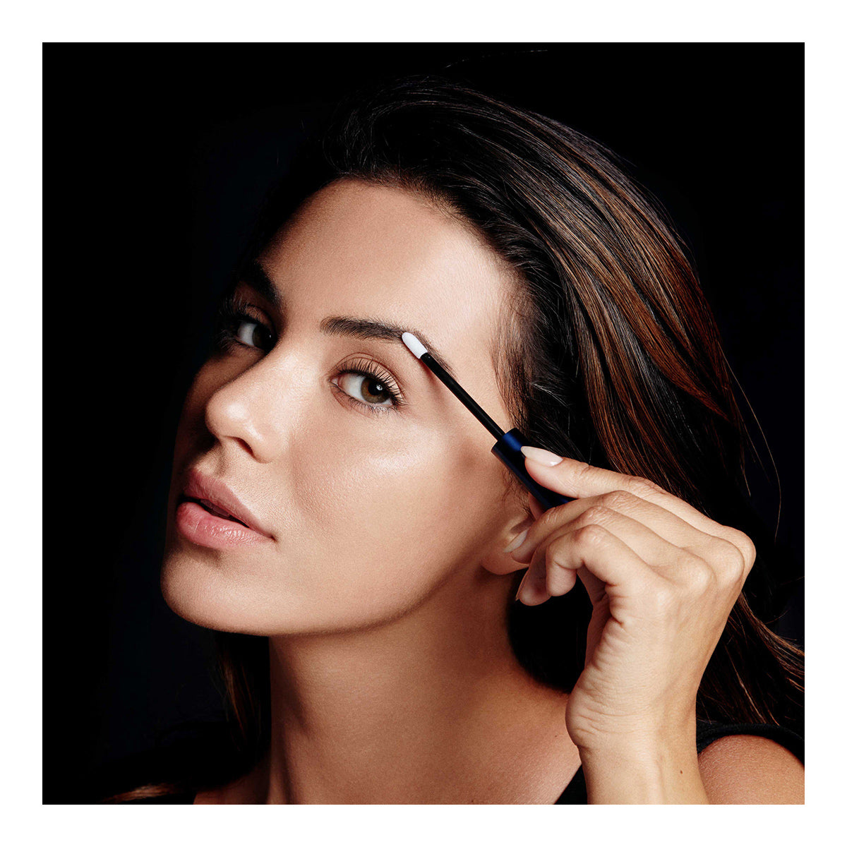 A woman with long dark hair uses a small brush to apply makeup to her eyebrows, looking focused and enhanced by RevitaLashs RevitaBrow Advanced Eyebrow Conditioner Serum, which combats brow aging. A dark background highlights her face.