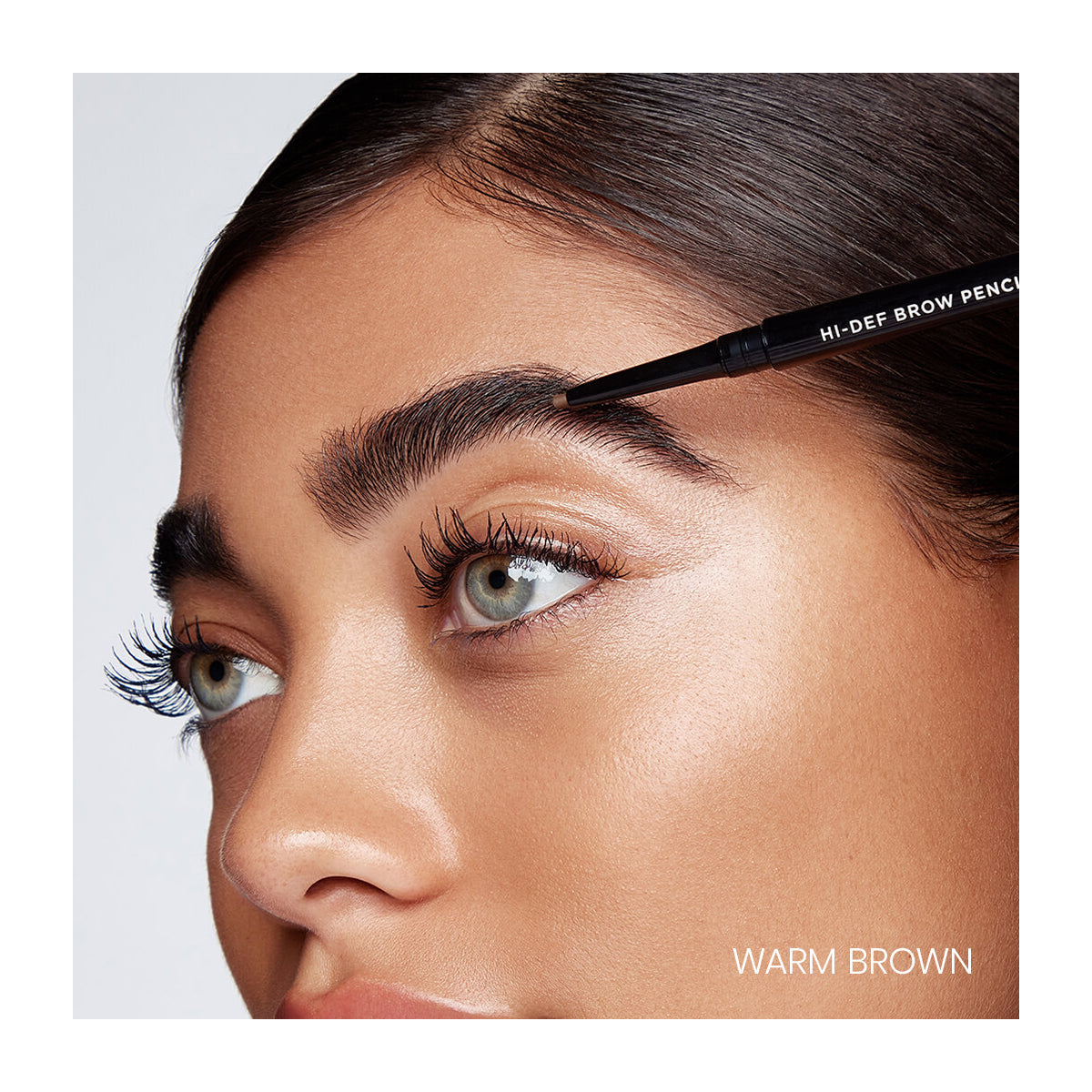 A person skillfully uses the long-wear, water-resistant RevitaLash Hi-Def Brow Pencil to enhance dark, thick brows. The image highlights well-groomed lashes and brows, with Warm Brown elegantly shown in the corner.