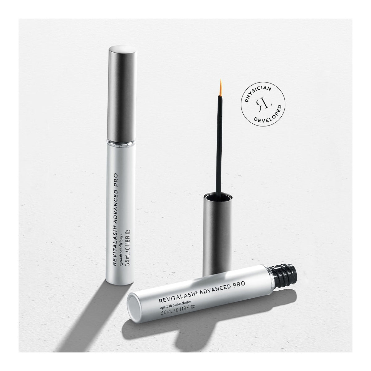 Two tubes of RevitaLash Advanced PRO Eyelash Conditioner and Enhancing Serum (3.5mL 6 Mo.Supply), one open with an applicator brush, the other closed, feature a sleek white design with silver caps on a gray surface beside a Physician Developed stamp, highlighting this innovative lash-enhancing formula.
