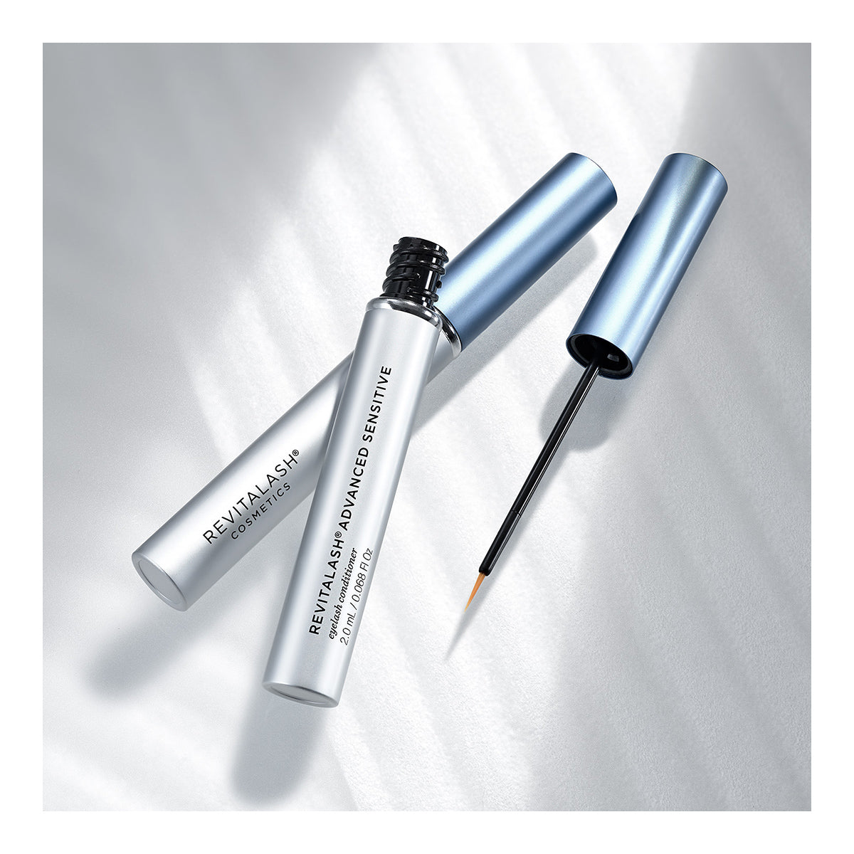 Two metallic blue tubes of RevitaLash Advanced Eyelash Conditioner and Enhancing Serum Sensitive (2.0mL 3 Mo.Supply) lie on a softly lit surface; one is open, showing the brush with Revitasome Technology, while the other stays closed and diagonal.