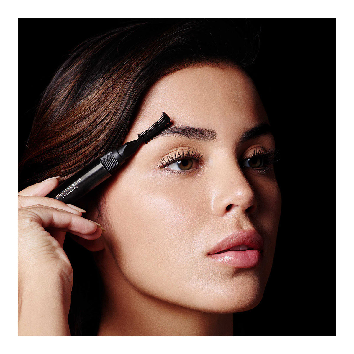 A person uses RevitaLash Hi-Def Brow Gel (3 Colors) and a precision styling brush for perfect lashes. They have long, dark hair, clear skin, and a neutral expression, all set against a black background that highlights their features.