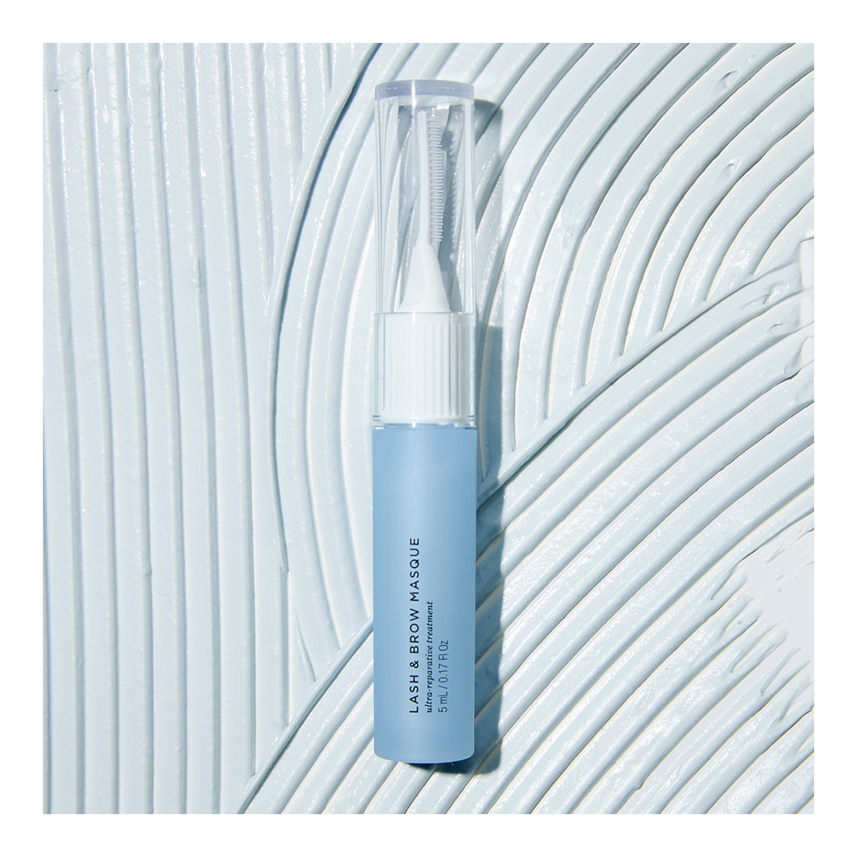 A blue tube of RevitaLash Lash and Brow Masque, designed for healthier lashes and brows, features a transparent cap. It rests elegantly on a textured white surface with sweeping curved lines.