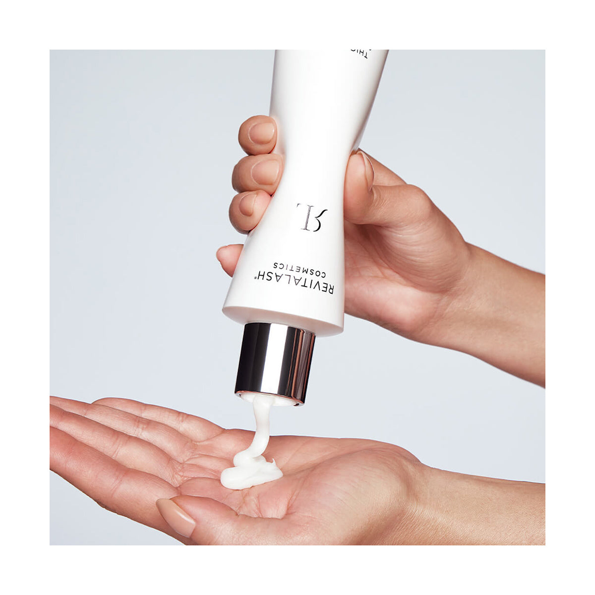 A person squeezes a white lotion from a RevitaLash Cosmetics Thickening Conditioner Scalp Therapy Formula tube into their palm, promoting scalp health. The tube has a white body with black text and a shiny silver cap, set against a plain, light background.
