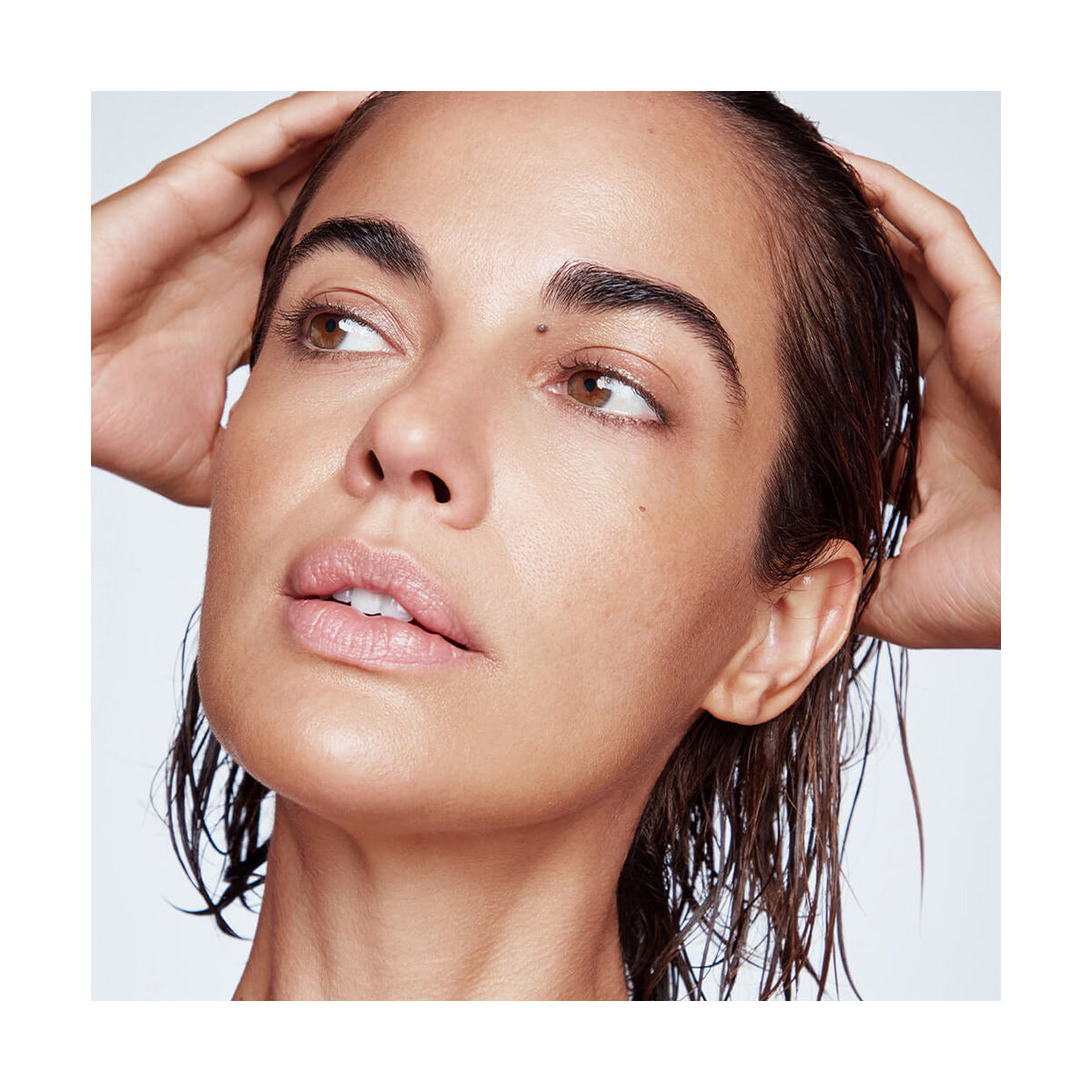 A person with wet hair treated with RevitaLash Cosmetics Thickening Conditioner Scalp Therapy Formula (Hypoallergenic, 250mL) gazes to the side, hands on head. The close-up highlights their smooth skin and natural makeup against a neutral backdrop that enhances their expression and complexion.