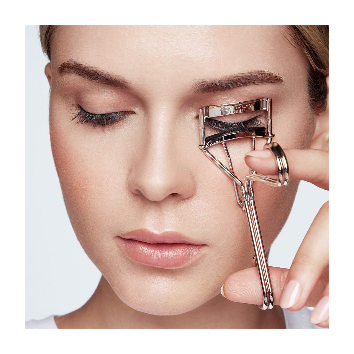 With eyes softly shut, someone uses the RevitaLash Signature Eyelash Curler, made of carbon steel with silicone pads, on their upper lashes. Their makeup includes foundation and a neutral lip color against a plain white background.