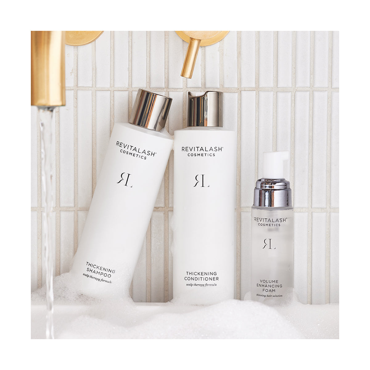 Three RevitaLash Volumizing Hair Collection Bundle bottles—Shampoo, Conditioner, and Foam—rest on a white tiled surface amidst water and bubbles. Infused with biotin, they transform the likely bathroom setting into a luxurious hair care sanctuary.
