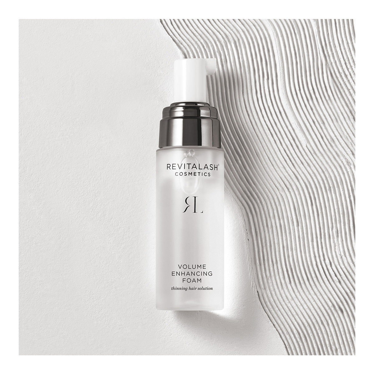 RevitaLash Volume Enhancing Foam for Hair (55mL) sits on a textured white surface. The sleek, semi-transparent bottle with a metallic pump features BioPeptin Complex, offering an ideal solution for thinning hair.