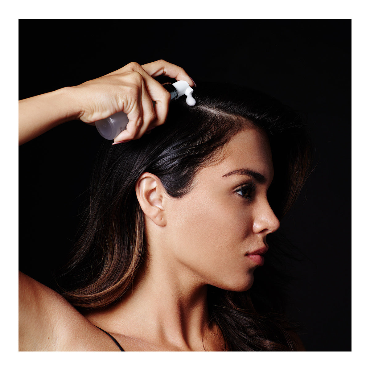 A person with dark hair expertly applies RevitaLash Volume Enhancing Foam for Hair (55mL) with BioPeptin Complex to their scalp, while the right hand holds the bottle against a black background, highlighting it as a solution for thinning hair.